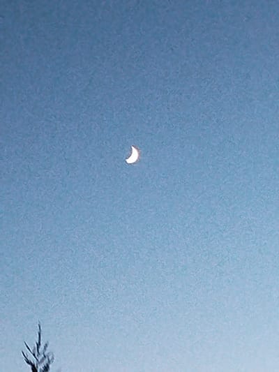 A crescent moon is visible in a light blue sky. A faint silhouette of a tree or branches is present at the bottom left of the image. The image is blurry and appears to have been taken at a distance, possibly with a low-resolution camera. The location is Kitzingen, Germany, as stated in the prompt.
