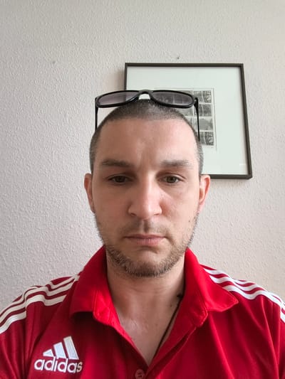 A portrait photo shows a man with short hair and stubble wearing a red polo shirt with white Adidas stripes on the shoulders. Sunglasses are perched on top of his head. He is looking directly at the camera. In the background, a framed picture hangs on a textured, light-colored wall. The scene appears to be indoors.