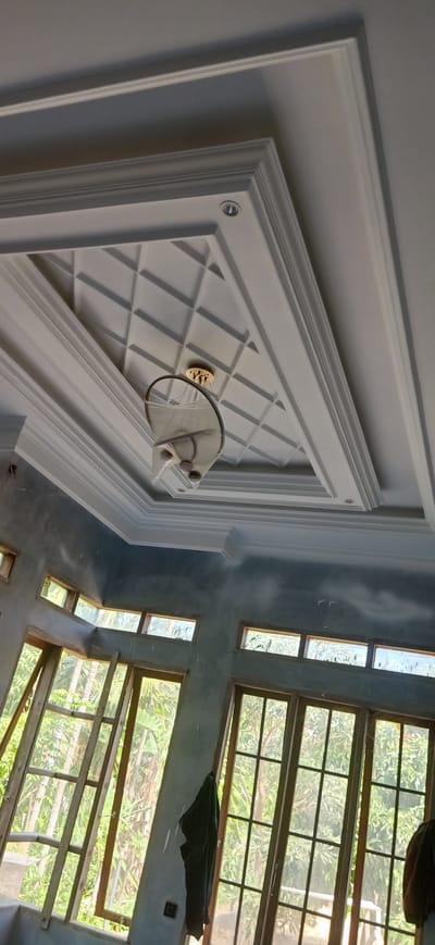 The image depicts the interior of a room under construction in Meunasahlhok, Indonesia. The ceiling features intricate white molding with a recessed central section containing a geometric pattern. A modern chandelier with a circular design and metallic accents hangs from the center of the ceiling. The walls appear to be freshly plastered and are a muted blue-gray color. Large windows with wooden frames are visible, offering views of lush green foliage and trees outside. Some of the windows are open, and a wooden ladder leans against one of them. A dark garment hangs on the wall near the windows, and a black electrical outlet is visible on the lower left wall. The overall impression is of a residential space in the process of being finished.