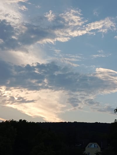 The image depicts a twilight scene over a wooded hillside in Baden, Austria. The sky is filled with a mix of light and dark clouds, with the setting sun casting a warm glow through a band of lighter clouds near the horizon. Silhouetted against the sky is a dark, tree-covered hillside. In the foreground, partially obscured by trees, is the upper portion of a yellow house with a distinctive arched window. The overall atmosphere is serene and natural.