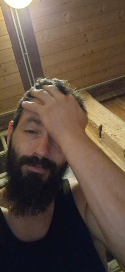 The image is a selfie of a man with a beard and dark curly hair, who is wearing a dark tank top. He has his right hand on his forehead. The background appears to be a rustic wooden interior, possibly a chalet or cabin, with visible wooden planks on the ceiling and a wooden beam. There is a light fixture visible in the upper left corner. The man's pose suggests he might be tired or stressed. The location is identified as Wattenwil, Switzerland.