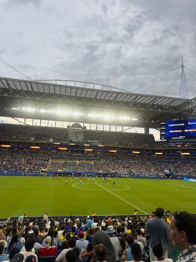 The image depicts a soccer match at Hard Rock Stadium in Miami Gardens, Florida, United States of America. A large crowd of spectators is seated in the stadium, filling the stands that surround the vibrant green soccer field. Several players are visible on the field, engaged in the game. Above the field, a large scoreboard displays information, including "SUBSTITUTION" and "FIFA WORLD CUP 2026". The stadium itself features a modern architectural design with a large, illuminated roof structure. The sky above is overcast with a cloudy, grayish hue. The overall scene captures the atmosphere of a professional soccer event.