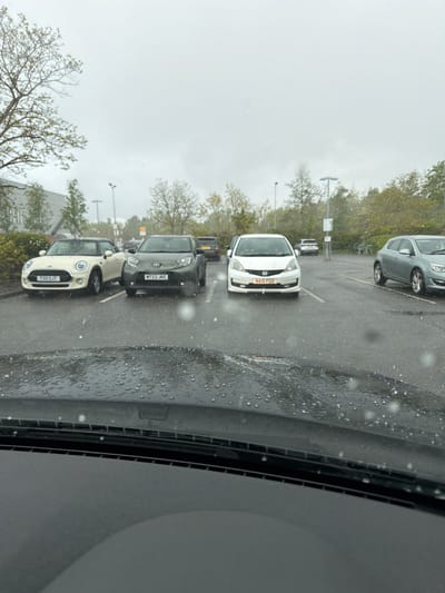 The image shows a view from inside a vehicle, possibly parked, at a car park in Pity Me, United Kingdom. The sky is overcast, and rain or hail is visible on the windshield. Several cars are parked in a row in the parking spaces. License plates visible include T33 SJT, KT23 JKL and N115 FGD. Trees and light poles are visible in the background. There is no evidence of people or activity other than parked cars.