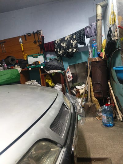 The image depicts a cluttered garage in Sychavka, Ukraine. A silver car occupies the foreground, with its hood partially visible. The garage interior is filled with various items, including tools hanging on a wooden board, a green tool case, black trash bags, clothing draped on a clothesline, a rusty metal barrel, a broom, and shovels. Several plastic bottles, including a large blue one, are also present. The walls are painted a light blue, and the floor is concrete. The overall impression is one of disarray and storage.