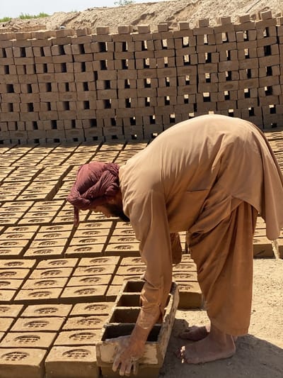 In Deh-E Bagh, Afghanistan, a man wearing a maroon head covering and a tan-colored outfit bends over a row of bricks. He is barefoot. His hands are immersed in a mold. Behind him, bricks are stacked in rows, filling the frame. The ground is dirt.