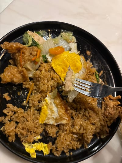 The image displays a close-up, top-down view of a partially consumed meal served on a round, glossy black plate, situated in Batuan, Indonesia. The plate contains a substantial quantity of brown-colored fried rice, interspersed with fragments of light green leafy vegetables, orange slivers, and darker green vegetable pieces. Two partially eaten fried eggs are visible, one with a distinct yellow yolk and browned white, and another fragmented with scattered yellow yolk remnants. On the left side of the plate are several pieces of golden-brown, textured meat, likely battered or glazed. A silver-colored metal fork is actively engaged in the food, piercing and slightly lifting a pale, striated piece of meat from the center-right section of the plate. The meal appears to be approximately half to two-thirds consumed. The black plate rests on a light-colored, smooth surface, suggestive of a table.