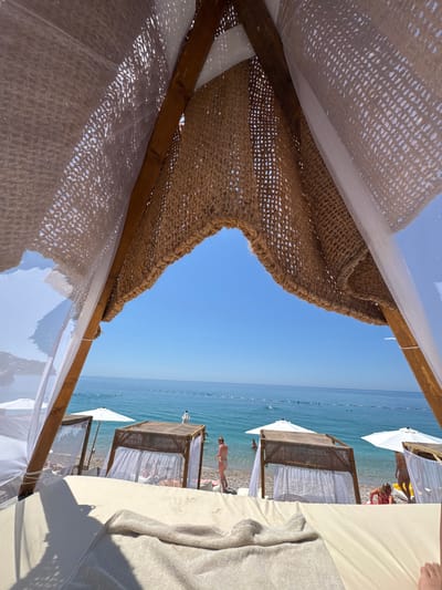 The image depicts a bright, sunny day at a beach in Vrba, Montenegro, viewed from within a beach cabana. In the immediate foreground, a light-colored, possibly beige, cushioned sunbed or daybed occupies the lower portion of the frame, with a matching towel draped over it and the edge of a foot visible in the bottom right corner. Above, the cabana's structure features rustic wooden beams supporting a roof composed of two layers: an inner sheer white mesh fabric and an outer coarse, woven brown material with a netting pattern.
Beyond the cabana, a pebble beach extends towards the water. Scattered along the shore are several other similar cabanas with white fabric canopies and white beach umbrellas providing shade. People are present on the beach and in the clear, calm, turquoise-to-deep-blue sea. On the right side of the beach, a woman in a light-colored bikini stands facing away from the viewer, near the water's edge. Another person in a red swimsuit is partially visible to her left, closer to the water. Further out in the water, a line of buoys marks a swimming area, with additional individuals swimming or wading. The horizon is clear, where the blue sea meets an expanse of bright, cloudless blue sky.