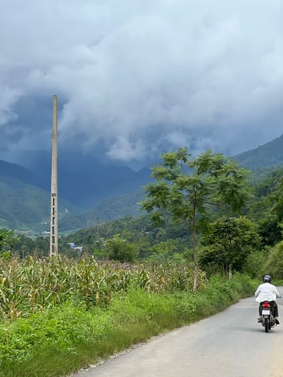 A person wearing a white shirt and a dark helmet rides a motorcycle down a paved road that curves to the right. The road is bordered by lush green vegetation, including a cornfield on the left and trees on the right. In the background, rolling green mountains are shrouded in a cloudy, dark sky. A tall, thin concrete pole with rectangular openings stands prominently in the cornfield, near the edge of the road. A small cluster of buildings is visible in the mid-ground between the cornfield and the mountains. The scene depicts a rural landscape in Lùng Tám Commune, Vietnam.
