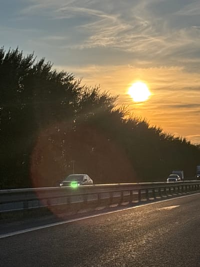 The image captures a sunset scene on a highway in Taymurzino, Russia. The sun, a bright, luminous orb, is low in the sky, casting a warm, golden glow across a band of thin, wispy clouds. The sun's light illuminates the sky with shades of yellow and orange, fading to a pale blue towards the left side of the frame.
In the foreground and to the right, a dark, dense line of trees borders the highway. The trees are silhouetted against the bright sky, their foliage appearing as a textured mass. The highway itself occupies the right side of the image, its asphalt surface textured and marked with faint white lines. A metal guardrail runs along the edge of the highway, reflecting the ambient light.
Two cars are visible on the highway. The first car, further down the road and closer to the trees, appears as a dark shape with some lights. The second car, nearer to the foreground and on the other side of the guardrail, is more distinct. It is a light-colored vehicle with its headlights on, casting a green glow. The overall atmosphere suggests an evening commute or travel during sunset. The lighting indicates it is likely late afternoon or early evening.