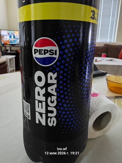 In the foreground, a large, black Pepsi Zero Sugar bottle dominates the frame. The bottle has a prominent yellow band across the top and features the Pepsi logo in a circular emblem. Vertical white text reads "ZERO SUGAR". The bottle appears condensation-covered. To the right of the bottle, a roll of white toilet paper is visible, with a small pink pattern on its surface. A yellow bowl is partially visible in the mid-ground. The background shows a blurred interior scene with a television, furniture, and a table with some items. The timestamp at the bottom indicates "12 июля 2026 г. 19:21". The location of Chal, Bulgaria is not discernible from the visual information in the image.