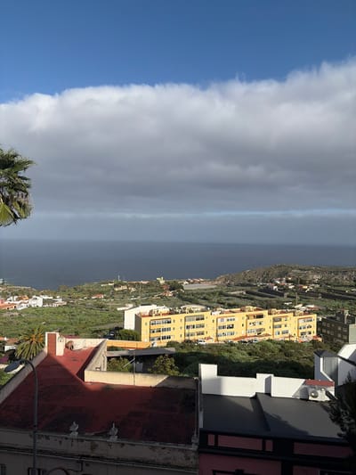 A high-angle, panoramic photograph depicts a coastal town in Icod de los Vinos, Spain, facing the Atlantic Ocean. The upper third of the frame shows a clear blue sky, transitioning downwards into a thick, horizontal band of white and grey stratocumulus clouds. Below the clouds, the ocean appears dark blue-grey, extending to the horizon.

The foreground features the rooftops of several buildings. On the lower left, a large, weathered reddish-brown tiled roof is partially shadowed. Adjacent to it, a flat-roofed building with light-colored walls and a terracotta chimney-like pipe is visible. To the right, a building with a black flat roof, white facade sections, and an exposed air conditioning unit is partially present. A portion of a palm tree frond is visible on the far left.

In the midground, an elongated, multi-story yellow apartment building with multiple windows and balconies stands prominently on a green, terraced hillside. Several white vehicles are parked on the green terrain near this building. Scattered across the surrounding green hillsides and towards the coast are numerous smaller white and light-colored residential structures, many with red or orange tiled roofs.

Further along the coastline, a small lighthouse structure is discernible on a headland to the left of a coastal indentation. The landscape consists of extensive green vegetation, including palm trees and what appear to be cultivated fields or natural scrub, descending towards the ocean. No individuals or specific activities are visible within the scene. The lighting suggests a clear day, with shadows indicating the sun is positioned to the left of the viewer.