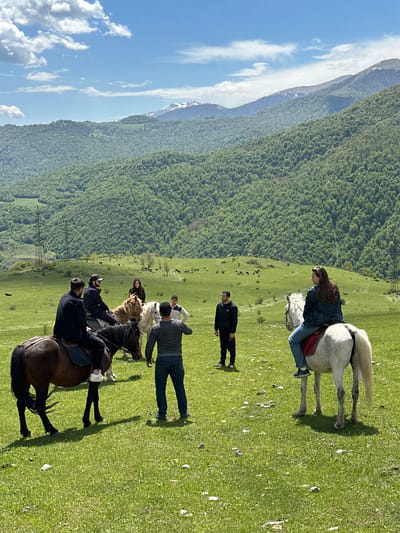 The image shows a group of people riding horses in a grassy field. Mountains and a forest are visible in the background, along with a blue sky with some clouds. Five people are on horseback, while one man is standing on the ground. The location is Teghut, Armenia.