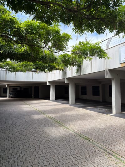 The image depicts an outdoor scene in La Clarté, France. The perspective is from below, looking up and to the side at a building and surrounding vegetation.
The primary subjects are a building with a modern, minimalist design and lush green trees and vines growing around and on it. The building appears to be multi-story, with visible concrete columns and awnings. Several dark, rectangular openings suggest windows or doorways on the upper levels. Below, a tiled walkway or parking area stretches into the distance.
The time of day appears to be daytime, with bright sunlight casting distinct shadows across the tiled surface, indicating a sunny or partly sunny day. The presence of shadows suggests a mid-morning or afternoon time. The weather seems pleasant, given the vibrant foliage.
Notable details include the texture of the tiled ground, which is made of small, uniform rectangular stones laid in a pattern. The vegetation is dense and healthy, with many leaves and some slender branches trailing. A metal pipe or conduit runs diagonally across the tiled area.
There is no discernible activity or event taking place; the scene appears still and possibly unoccupied at the moment the photo was taken. There is some faint, unreadable text on a plaque attached to the building on the far left.
The overall mood is one of tranquility and a blend of nature and architecture. The location cues, such as the architectural style and the foliage, are consistent with a Mediterranean or coastal European setting, which aligns with La Clarté, France.