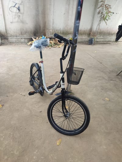 The image shows a small, white and black bicycle parked outdoors. The bicycle has a basket attached to the handlebars, and the seat is covered with a clear plastic bag. The ground is dirt, and there is a concrete wall in the background with some graffiti on it. A small tree is also visible in the background. The scene appears to be in an urban environment. This scene is located in Meunasahtimu, Indonesia.