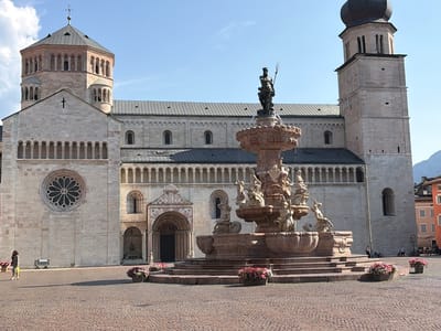 The image shows the Piazza Duomo in Trento, Italy, a public square dominated by the Trento Cathedral and a large fountain. The cathedral, a Romanesque basilica, features two distinct towers: one octagonal and adorned with arches, and another taller, more square tower topped with a dark dome. The facade of the cathedral is made of light-colored stone and includes a large rose window.
In the center of the square stands a prominent fountain, a multi-tiered structure adorned with sculptures. At its apex is a statue of a male figure holding a trident. The fountain is surrounded by a paved plaza, with flower pots containing colorful blossoms placed strategically around its base and along the edges of the square.
The scene is set on a clear day with a bright blue sky, suggesting it is daytime, likely late morning or early afternoon, given the sun's angle and the shadows cast. A few people are scattered across the square. One woman walks near the cathedral on the left, and several others are visible in the background near some buildings to the right. Bicycles are also present, parked near the people on the right.
The overall atmosphere is peaceful and serene, typical of a European city square. The architecture of the cathedral and the details of the fountain suggest a historical and culturally rich setting. The presence of people enjoying the public space indicates it is a lively, yet tranquil, urban environment. No visible text is present in the image.