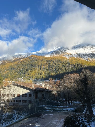 The image depicts a panoramic view of the La Thuile valley in Italy, observed from an elevated position. The upper portion of the frame features a bright blue sky with scattered cumulus clouds, some of which partially obscure the snow-capped mountain peaks in the background. Below the snowline, the mountains are densely covered with coniferous trees, predominantly showing vibrant golden-yellow autumn foliage, indicative of larch trees, with some darker green sections. A sharp diagonal shadow line extends across the middle slopes of the forested mountain.

In the mid-ground, a cluster of buildings characteristic of a mountain village is visible. A prominent, light-colored building with a multi-gabled terracotta-tiled roof and multiple chimneys occupies the left-center. Its walls are light grey, and several windows with dark wooden shutters are visible, some closed, some partially open. Portions of this building and its surroundings are cast in shadow. Further back, additional buildings with similar architectural styles are interspersed among trees.

The immediate foreground features a partially snow-covered environment. A tennis court, distinguished by its dark surface and white lines, is visible in the lower-center, with patches of snow and what appears to be ice. A net post is discernible on the left side of the court. Bare deciduous trees and bushes, also partially covered in snow, frame the tennis court and the buildings. No individuals are present, and no actions or interactions are depicted.