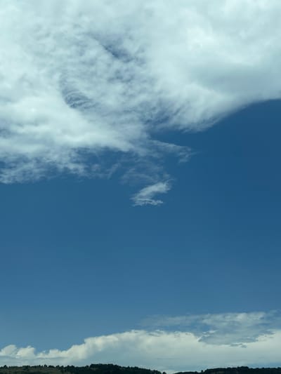 The image displays an expansive daytime sky over the distant landscape of Gorno Varshilo, Bulgaria. The upper third of the frame is dominated by a mass of white and light grey clouds, with a notable textured, swirling formation present in the upper-middle-left area. A sharp, diagonal edge of a cloud bank cuts across the upper-right quadrant, transitioning to a clear, deep blue sky that occupies the majority of the central and mid-right portions of the image. Along the lower horizon, a band of white and grey cumulus clouds is visible, resting just above a distant, dark green landscape characterized by undulating hills covered in dense foliage. No people, animals, man-made structures, or specific objects are discernible within the frame. The lighting indicates a bright, sunny day.