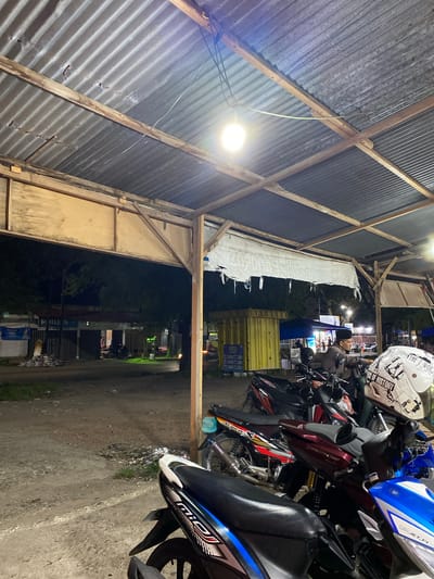 The image depicts an outdoor scene at night, likely in the city of Payatieng, Indonesia, with a focus on a collection of parked motorcycles under a corrugated metal roof supported by wooden beams. A single light bulb hangs from the ceiling, illuminating the immediate area. Several motorcycles are visible in the foreground, some with visible branding such as "XMILE-FI" on a blue and white one. A white helmet with text and graphics, including "FREE OF HISTORY" and "THE 7 WORDS", rests on top of a motorcycle. In the background, there are structures that appear to be shops or stalls, with some neon lighting. A man, wearing a dark shirt and a peci (a type of cap), is seated on one of the motorcycles, looking forward. The ground is a mixture of dirt and gravel, with some scattered debris. The overall impression is that of a parking area or a roadside stop, possibly associated with local businesses or a transportation hub.