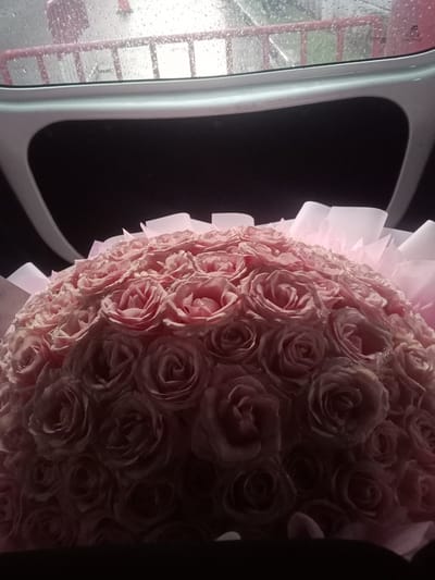 A bouquet of light pink roses is positioned in the foreground of the image. The roses are densely packed and appear to be slightly wet, with small water droplets visible on the petals. The bouquet is wrapped in translucent pink paper. Above the roses, a white, curved structure is visible, and beyond that, a window with raindrops on it. Through the rainy window, a pink railing can be seen. The background is dark. The image appears to have been taken inside a vehicle or enclosed space, possibly looking out through a sunroof or rear window. The location is described as Pakulonan Barat, Indonesia.