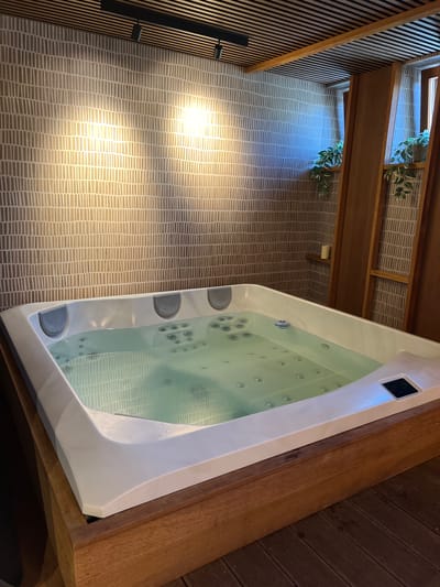 The image shows a well-lit, modern spa area, likely within a hotel or private residence in Mengusovce, Slovakia. Dominating the foreground is a large, white, built-in jacuzzi filled with clear, slightly rippling water. The jacuzzi has multiple jets visible beneath the surface and a digital control panel on its right side. The tub is surrounded by a wooden structure, and the floor is made of dark wooden planks. The background features a textured, neutral-toned wallpaper with a repeating vertical line pattern. Above the wallpaper, a slatted wooden ceiling with track lighting is visible. To the right, wooden panels create a partition, revealing glimpses of windows with greenery, possibly potted plants, on ledges. A small, unlit candle sits on a shelf within this wooden structure. The overall impression is one of relaxation and contemporary design.