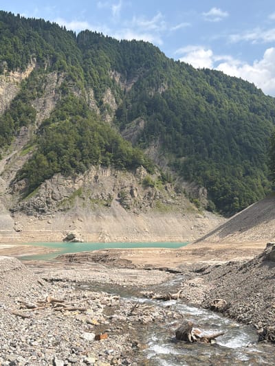 The image shows a mountainous landscape with a body of turquoise water in the middle ground, likely a reservoir or lake. The surrounding slopes are covered in dense green trees, with exposed rocky sections on the mountainside. In the foreground, a rocky riverbed with a shallow stream flows towards the water. The scene appears to be in a natural, undeveloped area, consistent with the mountainous terrain of Ampezzo, Italy. There are no people or specific man-made structures visible that would pinpoint a precise location within the city. The weather appears to be clear with some clouds in the sky.