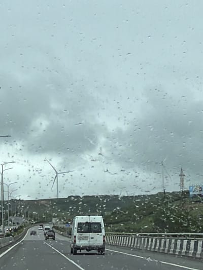 The image, taken through a rain-streaked window, depicts a view of a highway and landscape near Otarsheni, Georgia. Wind turbines are visible in the distance. There is a white van traveling on the right side of the road with other vehicles behind it. The side of the highway features a concrete barrier with black and white stripes. A green, hilly area borders the left side of the highway, with additional infrastructure, including streetlights, visible in the foreground. The sky is overcast.