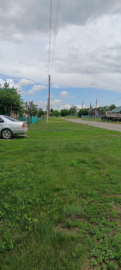 The image depicts a street scene in the village of Kamennoye, Russia, on a cloudy day. A silver sedan is parked on the left side of the frame, next to a grassy area with a teal-colored fence. The street is unpaved and curves to the right, leading into the distance where more houses and trees are visible. Power lines run overhead, supported by wooden poles. A tractor and trailer are parked on the right side of the road. The overall impression is one of a quiet rural setting.