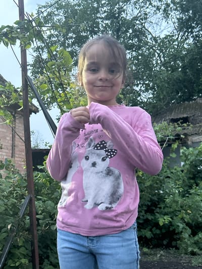 A young girl with dark hair and bangs stands outdoors, facing the camera. She is wearing a pink long-sleeved shirt with a graphic of a rabbit wearing a bow tie, and blue denim jeans. Her hands are clasped in front of her chest. She is positioned in front of a backdrop of dense green foliage. In the background, to the left, a brick structure with an angled roof is visible, and to the right, another structure with a similar roofline and some surrounding greenery. The scene appears to be in a residential yard, possibly in the city of Prokhladny, Russia, given the common architectural styles and vegetation. The lighting suggests it is late afternoon or early evening.