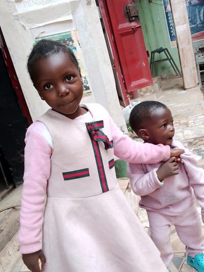 This image captures two young children standing outside, possibly in an urban setting in Mbar, Nigeria, given the context. The primary subject is the older girl in the foreground, who is looking directly at the camera with a gentle, curious expression. She is wearing a light pink dress with a textured herringbone pattern and a contrasting pink sweater with fluffy sleeves. The dress is adorned with a green and red striped ribbon at the chest and a matching ribbon detail on the skirt.
To her right, a younger child, likely a toddler, is reaching around her, their face turned away from the camera and looking to the right. This child is dressed in a light pink ribbed tracksuit. Both children appear to be in good spirits.
The background shows an outdoor environment with weathered walls and a red metal door. There are posters and signs on the wall, some of which are partially visible and contain text. One poster advertises the "COLLEGE OF EDUCATION GINDIRI, PLATEAU STATE" and mentions "THE REGISTRAR" and a website "Jeg.edu.ng". Another visible text fragment reads "Product" and "11338064". The ground is paved with tiles. The lighting suggests it is daytime, and the scene appears to be a street or a courtyard.