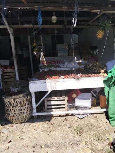 A bustling fruit stand displays an array of produce. Rambutans dominate the forefront, overflowing from a large woven basket and lining the white table. Dragon fruits are stacked in wooden crates to the side. Behind, other fruits like apples, and what appears to be garlic bulbs are arranged. A pineapple hangs near the edge of the roof, adding to the vibrant display. Boxes are stacked haphazardly beneath the table, and fallen fruit litters the ground. A green cloth drapes on the right side of the table.