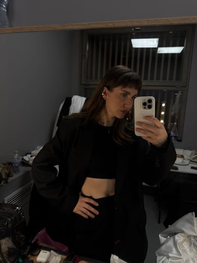 The image shows a young woman with brown hair and bangs, wearing a black crop top and an oversized black blazer. She is holding a white smartphone in her right hand, taking a selfie in a mirror. Her left hand is on her hip. The background appears to be a backstage dressing room, with clothing racks, a table with various items, and a mirror reflecting the room. The environment is dimly lit, with vertical blinds visible in the reflection. The woman is wearing a small silver necklace and a silver earring. There are no other people clearly visible in the image. The location is not specified in the prompt, and therefore cannot be provided.
