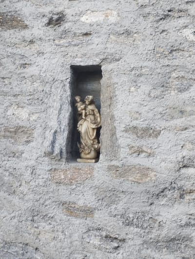 A statue of a woman holding a child is displayed in a small niche carved into a rough, grey stone wall. The statue is made of a light-colored material, possibly stone or plaster, and appears to be weathered. The woman is depicted with flowing robes, and the child is held in her arms. The statue is placed on a small pedestal that rests on the bottom of the niche. The wall itself is textured with uneven stones and mortar, suggesting it is an outdoor structure, possibly part of a building or a monument. The lighting is soft, indicating daylight, but the overall impression is one of stillness and quiet contemplation. The setting appears to be a historical or religious site, given the nature of the statue and its placement. There is no visible text in the image.
