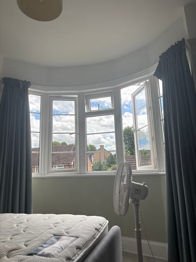 A bay window with white frames dominates the scene, showcasing a clear sky with scattered clouds and glimpses of neighborhood rooftops. Gray curtains frame the top and bottom of the window. A white stand fan is positioned to the right, partially obstructing the view. To the right, a mattress sits upright. A lamp is visible in the lower left corner, attached to the ceiling. The walls are painted in a light neutral tone.