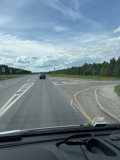 This is a vertical, eye-level shot of a highway in Stolbishche, Russia. The road is asphalt and has white lane markings. There are trees lining the left side of the road, and a bright blue sky with white clouds fills the top half of the image. A black car is driving on the highway, and it is the main subject of the image. The time of day is daytime, as indicated by the bright sky and sunlight. The weather appears to be fair. A yellow line is visible on the right side of the road. There is a signpost with traffic signs visible in the distance. The photo appears to be taken from a vehicle.