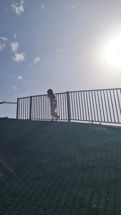 A young person in pink clothing and white shoes is climbing on a playground fence at an angle on a sunny day. The location appears to be an outdoor playground with a dark green rubberized surface. A fence with horizontal bars is visible. Part of a light blue sky with a few scattered clouds is visible above, with the sun at the top of the frame. The image appears to be taken in Kiryat Gat, Israel.