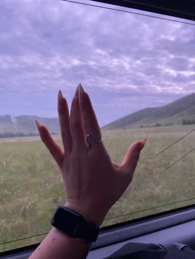 A hand with long, pointed, light-colored fingernails rests against a window. The hand is adorned with a thin, silver ring that has a wavy design. On the wrist, a black fitness tracker is visible. Through the window, a blurred landscape of green fields and rolling hills under a cloudy, purple sky can be seen. The scene suggests the perspective of someone traveling, possibly by car or train, through a rural area near Petrovskoye, Russia.