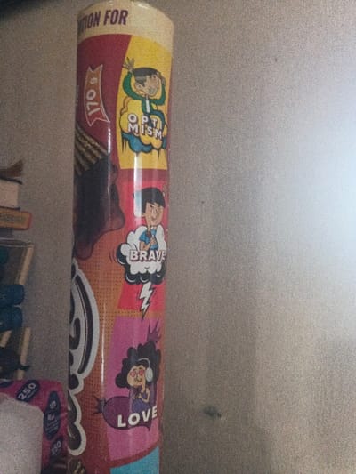 The image depicts a vertical, cylindrical object, likely a poster roll or column, positioned on the left side of the frame against a light-colored, slightly textured wall. This scene is located in Dogsenang Gorphoog, Bhutan.
The cylindrical object is wrapped in a multi-paneled graphic design in a comic book or pop art style. From top to bottom, visible panels include:
1. A yellow panel featuring a cartoon figure of a person with dark hair and raised arms, smiling, above a cloud graphic containing the word "OPTIMISM".
2. A reddish-orange panel showing a cartoon figure of a person with dark hair in a light blue shirt, with one arm raised, above a cloud graphic containing the word "BRAVE". A white lightning bolt graphic is present below this.
3. A pink panel displaying a cartoon figure of a person with curly dark hair wearing headphones, holding a purple heart, with heart-shaped eyes, above the word "LOVE".
To the immediate left of these panels, a dark red section of the cylinder is visible, with the text "170 G" oriented vertically. A light blue section is visible at the very bottom of the cylinder.
In the blurred background on the far left, partially obscured objects are stacked, possibly books or other items, with a white item marked "250" visible at the bottom. The plain wall on the right occupies approximately two-thirds of the image, appearing brighter than the left side, suggesting a light source from the right. The overall image has a grainy texture.