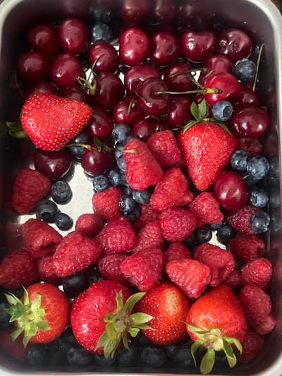 The image presents a close-up, overhead view of an assortment of fresh berries and cherries contained within a rectangular, metallic vessel, likely stainless steel. The container is filled with a dense accumulation of fruits, appearing recently washed as evidenced by moisture and reflective surfaces on both the fruit and the container's interior.
Prominently featured are numerous bright red strawberries, some displaying intact green calyxes, distributed throughout the lower and middle portions of the composition. Dark red, glossy cherries, many with their green stems still attached, are concentrated towards the upper and mid-sections. Vibrantly red, textured raspberries are interspersed, forming significant clusters in the mid-ground. Small, dark blue blueberries are scattered among the other fruits, providing textural and color contrast.
No individuals or specific actions are depicted. The immediate environment is confined to the interior of the container. The image provides no discernible information regarding its geographical location, including the city of Herne, Germany.