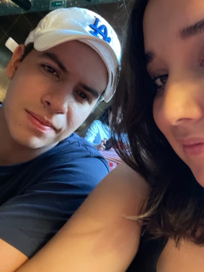 A close-up, selfie-style photograph captures two individuals in an indoor establishment. The primary subject is a young adult male, positioned on the left, looking directly at the camera with a slight smile. He has dark hair, dark eyes, and a light complexion, and wears a white baseball cap with a prominent blue "LA" logo on the front. He is dressed in a dark blue short-sleeved t-shirt, with his left arm folded across his chest. Partially visible on the right is a second individual, a female, whose right side of her face (including her nose, mouth, and a portion of her right eye) and upper arm are in the foreground, indicating she is likely holding the camera. She has dark hair and a light complexion, and appears to be smiling faintly. In the background, out of focus, another person wearing a light-colored striped shirt is seated at a table covered with a red and white striped tablecloth. A white "EXIT" sign with red lettering is visible on a wall in the upper left background. The lighting is warm and indirect. The image does not contain specific visual information to confirm the location in Boca del Río, Mexico.