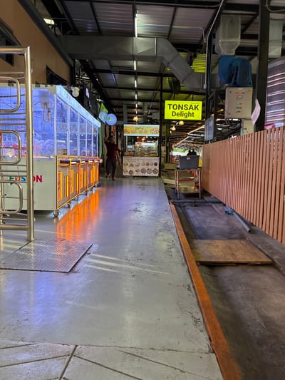 The image depicts an indoor market or arcade area with a concrete floor. In the foreground, a metal grate and a metal plate are visible. To the left, a large arcade claw machine is illuminated with blue and pink lights, displaying stuffed animals inside. The machine has the word "FUSION" printed on it. To the right of the claw machine, a food stall is set up with various items displayed. A neon yellow sign reading "TONSAK Delight" is prominently displayed overhead, along with Thai text. A man in a dark red shirt and grey pants is walking away from the camera towards the food stall. Further down the walkway, a wooden fence runs along the right side of the image. In the background, there are ventilation systems and lighting fixtures on the ceiling. The overall scene suggests a public commercial space, possibly a night market or entertainment venue, located within the city of Nonthaburi, Thailand.