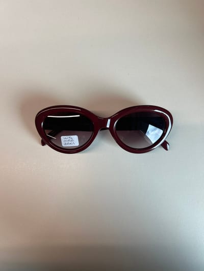 A single pair of dark red, oval-framed sunglasses with dark gradient lenses rests on a plain, light beige surface. The frames are glossy, and the lenses reflect ambient light. A small, rectangular white sticker is affixed to the upper portion of the left lens (viewer's left), displaying handwritten text in blue ink: "W13" above "metal detail." The sunglasses are positioned horizontally in the center of the frame, with the temples folded inward. A subtle shadow is cast beneath the object, suggesting uniform overhead lighting. No individuals, actions, or specific interactions are depicted. The environment is entirely generic, providing no identifiable features to determine a specific location within the city of Garston, United Kingdom, or any other geographical area.