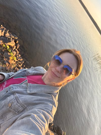 A woman, appearing to be in her 30s, with blonde hair and wearing sunglasses, a pink t-shirt, and a denim jacket, takes a selfie while standing by a body of water. The environment appears to be a natural setting, with rocks and vegetation visible along the shoreline. The water is calm and reflects the light of what appears to be either sunrise or sunset. The overall impression is of a casual, outdoor moment. The image was taken in Shuiskaya Chupa, Russia.