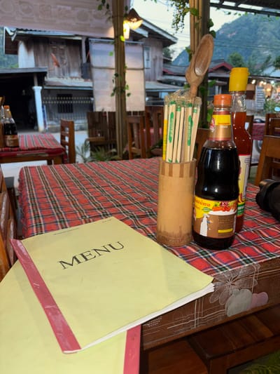 The image depicts an unoccupied dining table at an outdoor or semi-outdoor restaurant in Nongkhiao-Nua, Laos. The table is covered with a red and blue plaid tablecloth over a wooden surface. On the table, two yellow menus with pink bound edges are present, with the top menu displaying "MENU" in black lettering. A cylindrical bamboo holder stands beside the menus, containing multiple individually plastic-wrapped chopsticks and a wooden spoon-like utensil. To the right of the utensil holder are two condiment bottles: a large, dark brown bottle labeled "SEASONING SAUCE" with a white label featuring a chef graphic, and a taller, slender bottle with a yellow cap containing a reddish liquid. A dark object, possibly a camera lens, is partially visible on the far right edge of the table.
In the mid-ground, additional wooden tables with similar plaid tablecloths and wooden chairs are visible, suggesting more seating within the establishment. Support pillars for the restaurant structure are present, adorned with artificial green vines and a hanging light fixture emitting a warm, yellowish glow. The background reveals an older, two-story wooden building with a corrugated metal roof and a satellite dish attached to its facade. Beyond this building, lush green mountains rise under an overcast or twilight sky, indicating a mountainous setting. No individuals are present or engaged in any actions within the frame.