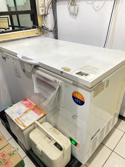 This media file shows the interior of a large, white freezer, likely in a medical or laboratory setting in Margoyoso, Indonesia. The freezer is filled with various items, including a white plastic container labeled "OneMed" and several boxes wrapped in clear plastic and brown paper, some with red and blue markings. One box appears to be a medical shipment.
Prominently displayed on the side of the freezer is a circular sticker indicating temperature ranges: "+43°C" in orange and "+5°C" in blue. There is also a digital display on the freezer's control panel showing "14.5", presumably a temperature reading.
The freezer itself is a chest-style model with a white lid and a silver handle. The interior walls are also white. On the exterior of the freezer, there are several labels and stickers, including one that reads "R134a," which is a type of refrigerant.
Additional details in the background suggest the location is a somewhat cluttered storage area. Wires and electrical outlets are visible on a white wall, and a metal rack is present. The overall impression is one of a functional, utilitarian space focused on storage and preservation. The image is well-lit, suggesting it was taken during the daytime. No people are visible in the frame.