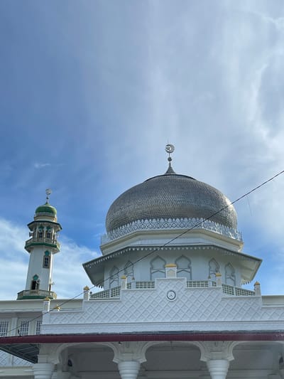 The original image, oriented sideways (rotated 90 degrees clockwise), presents an architectural view of the Mesjid Reubee mosque in Indonesia, captured from a low angle against a bright, partially cloudy blue sky.
The most prominent feature is a large, metallic silver-colored dome on the main mosque building, covered in a detailed, scale-like pattern that reflects the light. The base of the dome is adorned with white, intricate latticework and decorative trim. To the right of the dome (when viewed upright), the mosque's white facade features deeply carved geometric patterns and arched window-like structures with golden accent spires. A small, round clock face is visible on one of the patterned sections of the wall, though its numbers are not discernible.
Further down and to the left of the main dome (when viewed upright), a tall, slender minaret rises towards the sky. The minaret is predominantly white, featuring distinct green and gold-colored sections, multiple balconies with railings, and arched window details. Both the main dome and the minaret are topped with crescent moon and star finials, symbolic elements in Islamic architecture. Thin black lines, possibly power lines or wires, stretch diagonally across the sky in the upper portion of the image. The bright daylight suggests it is daytime, with soft shadows highlighting the architectural details. No people or specific activities are visible in the still scene.