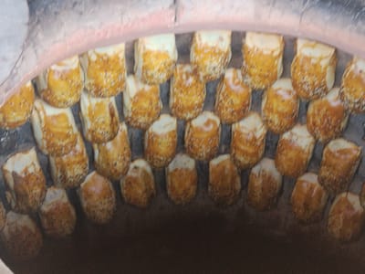 The image shows a close-up, slightly blurred view from above into a tandoor oven. The oven walls are visible at the top and sides, constructed of brick and lined with a reddish-brown material. Inside the oven, numerous round, bread-like items are arranged in rows, attached to the curved inner wall. These items are golden brown with darker brown, almost charred, areas, and appear to be sprinkled with sesame seeds. They are cylindrical in shape, tapering slightly towards the top, with a flat top surface. The lighting suggests the interior of a hot oven, with some highlights and shadows indicating the curvature of the items and the oven. The overall impression is of freshly baked goods within a traditional tandoor oven, likely in Ala-Buka, Kyrgyzstan, where such ovens are commonly used for baking bread and other dough-based foods.