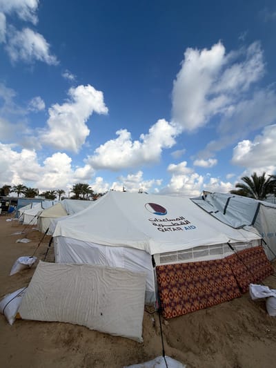 This image depicts a temporary settlement or encampment located in a-Zawayda, Palestinian Territory. The foreground is dominated by a large white relief tent with "QATAR AID" printed on its side in English and Arabic ("المساعدات القطرية"), accompanied by the Qatar national flag emblem. The tent is secured with visible guy lines and weighted down by white sacks, presumably filled with sand, around its base. Leaning against the tent's exterior, on the left, is a plain, light-colored mattress, while two patterned mattresses with red, orange, and brown geometric designs lean against the right side.

To the left and slightly behind the main tent, a row of several similar white and light-colored tents extends into the distance, indicating a larger camp structure. Another tent, partially covered with what appears to be a grey or blue tarp, is visible immediately to the right of the main tent. The ground beneath the tents is sandy and barren. In the background, numerous palm trees are visible, characteristic of the region. The sky above is a bright blue, dotted with scattered white cumulus clouds. No individuals or direct actions are depicted.