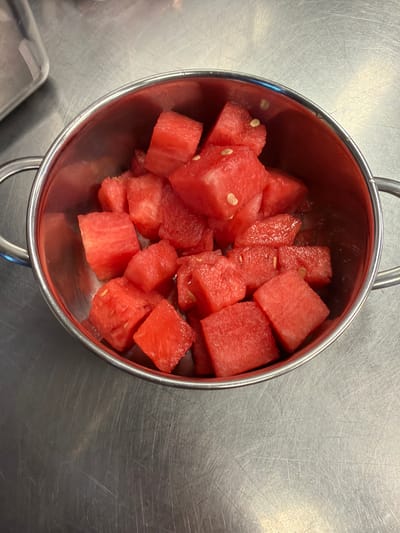 A close-up, top-down view shows a metallic bowl filled with diced watermelon. The watermelon pieces are bright red and contain visible seeds. The bowl has two handles and rests on a brushed metal surface. The background is out of focus and appears to be a kitchen or food preparation area. There are no people or animals visible in the image. The scene is static and appears to be a still life. The location in Rust, Germany, is not visually identifiable from the image.