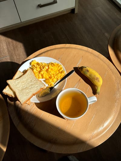 A breakfast arrangement rests on a wooden tray. A plate holds scrambled eggs and white bread toast, accompanied by a black knife. Adjacent to the plate is a white mug filled with a yellow liquid. A slightly bruised banana completes the meal. The scene is illuminated by bright sunlight, casting harsh shadows across the tray and surrounding surface.
