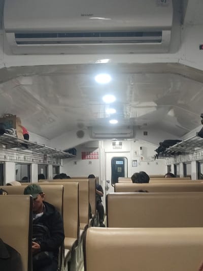 The image shows the interior of a train car. There is a white air conditioner unit with the brand name "SHARP" printed on it, mounted on the ceiling. The ceiling is white and there are multiple overhead fluorescent lights. The train car is sparsely populated. There are seats arranged in rows on each side of the train car, which are covered in brown faux leather. Several people are sitting on the seats. One person is visible, a man with a dark green baseball cap. He is wearing a black jacket and appears to be looking at his phone. The back of the seats in front of him are empty. The interior walls of the train car are white and there are luggage racks above the seats. There is a door in the center of the train car. The train car is in a train station in Kecamatan Gempol, Indonesia, which is located at latitude: -7.568, longitude: 112.712.